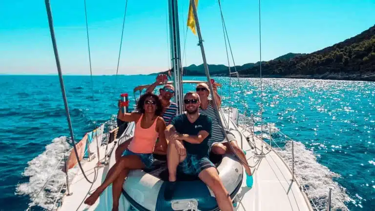 Vier Freunde genießen einen sonnigen Tag auf einer Segelyacht und winken in die Kamera, während sie vom klaren blauen Himmel und dem glitzernden Meerwasser umgeben sind.