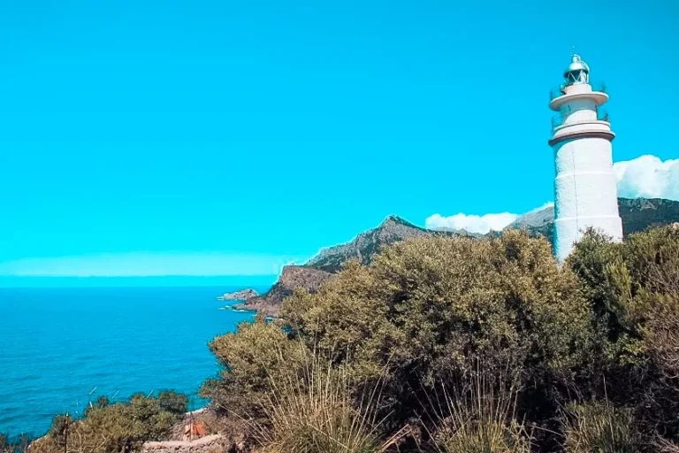 Ein weißer Leuchtturm steht in einer üppigen Küstenlandschaft mit Blick auf ein ruhiges blaues Meer und in der Ferne sind unter einem klaren Himmel Segelboote zu sehen.