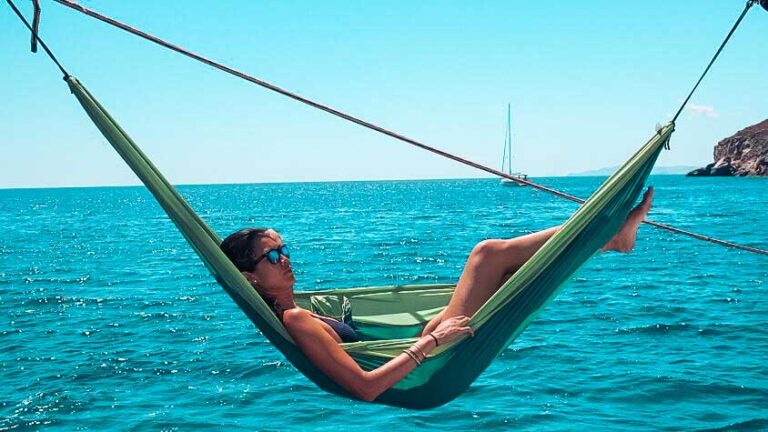 Eine Person entspannt sich in einer grünen Hängematte über dem klaren, blauen Meer unter einem strahlend sonnigen Himmel. In der Ferne sind Hügel und im Hintergrund eine Segelyacht zu sehen.