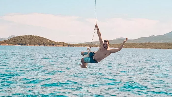 Ein Mann schwingt während seines Segelurlaubs mit einem Lächeln im Gesicht an einem Seil über einem klaren, blauen Meer, mit Bergen im Hintergrund und einem strahlenden Himmel.