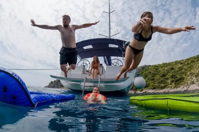 Zwei Menschen springen freudig von einer schwimmenden Plattform ins Meer, im Hintergrund sitzt eine Frau auf einer Segelyacht nahe einer üppigen Küste.