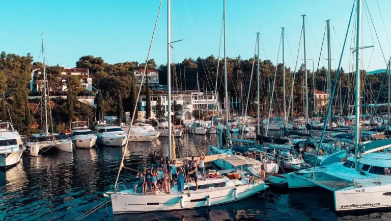 Eine Gruppe von Menschen auf einer Segelyacht ist bereit zur Abfahrt, umgeben von anderen angedockten Segelbooten in einem lebhaften Yachthafen mit dicht bewaldeter Küste im Hintergrund.