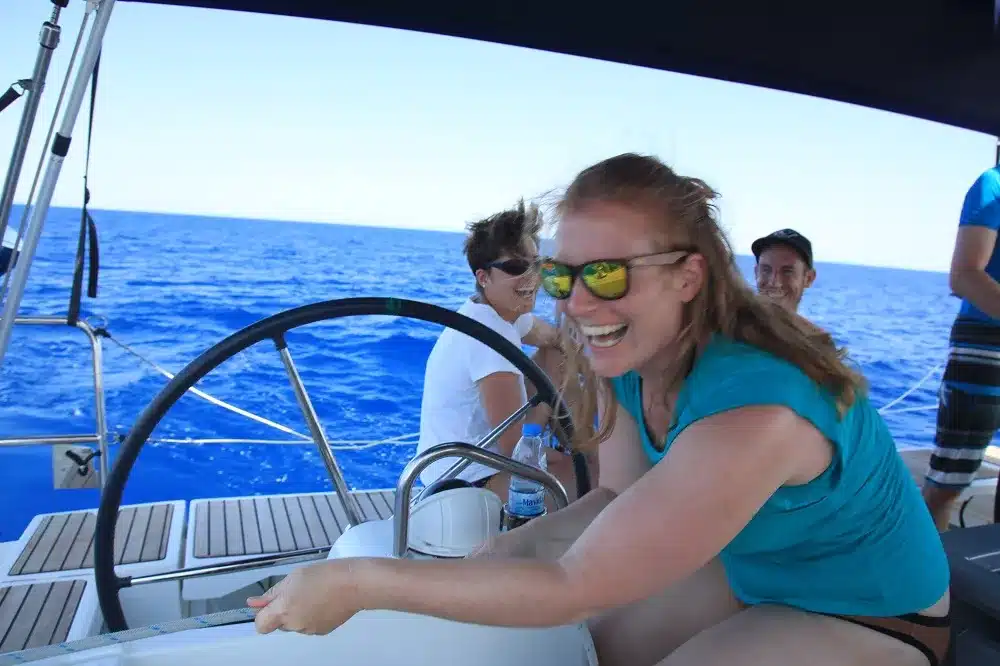 Eine fröhliche Frau mit roten Haaren steuert einen Katamaran. Sie trägt ein blaues Tanktop und eine verspiegelte Sonnenbrille, im Hintergrund ist der klare blaue Himmel und das Meer zu sehen. Hinter ihr sind weitere Passagiere zu sehen.