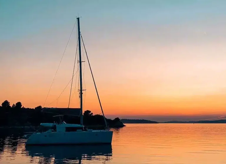 Eine Segelyacht auf ruhigen Gewässern während eines Sonnenuntergangs mit leuchtenden Orange- und Rosatönen am Himmel, die sich im Wasser nahe der Silhouette einer bewaldeten Küste spiegeln.