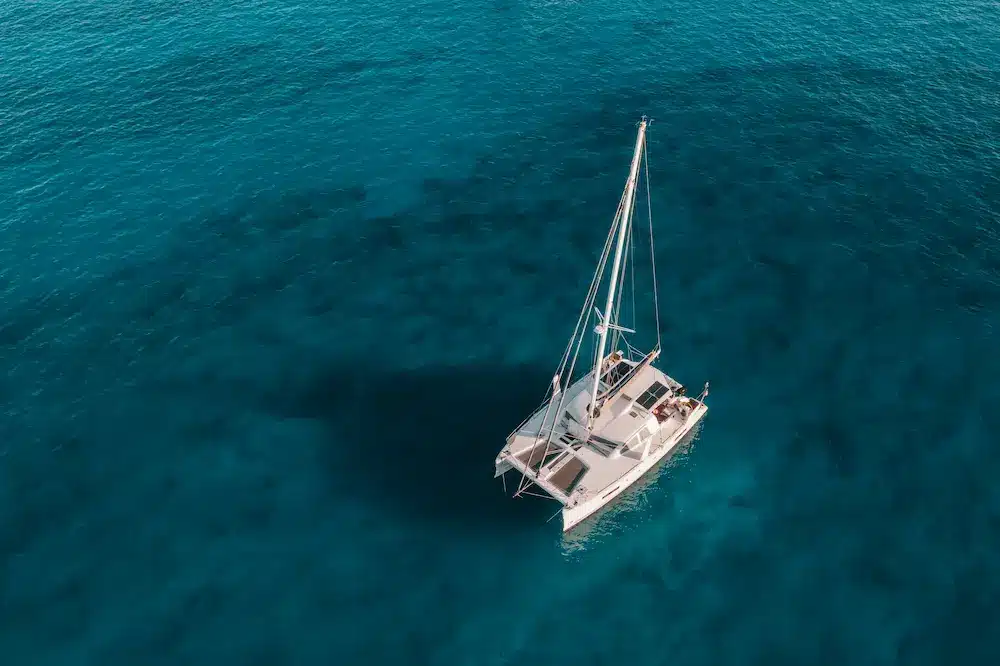 Luftaufnahme eines weißen Segelboots auf einem ruhigen blauen Ozean. Die Aufnahme zeigt einen deutlichen Kontrast zwischen dem Boot und dem tiefblauen Wasser, das es während eines Segelurlaubs umgibt.