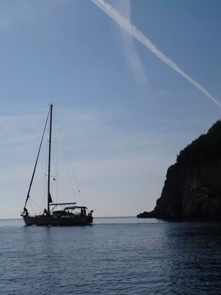 Eine Segelyacht liegt vor der Küste in der Nähe einer felsigen Küste unter einem klaren blauen Himmel. Am Himmel ist deutlich ein Kondensstreifen sichtbar.