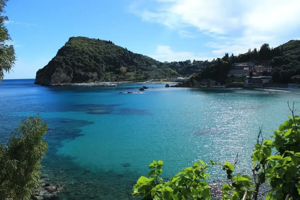 A peaceful seascape with calm blue-green waters beside a lush green peninsula under a bright blue sky. Coastal buildings and a sailing yacht are nestled among trees on the right side.