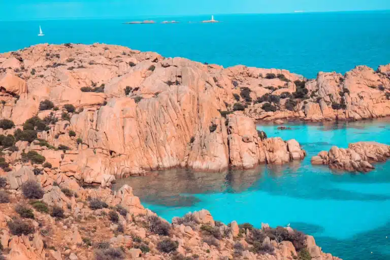 Luftaufnahme einer felsigen Küste mit türkisfarbenem Wasser, mit markanten Formationen aus rosa Granitfelsen und mehreren Segelbooten, darunter Segelyachten, im fernen Meer.