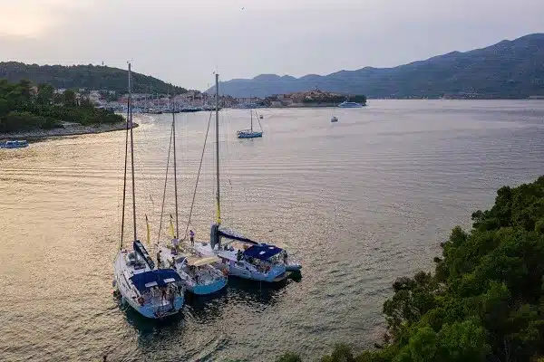Luftaufnahme von drei Segelyachten, die dicht beieinander in einer ruhigen Bucht vor Anker liegen, mit einer malerischen Küstenstadt und Hügeln im Hintergrund in der Abenddämmerung.