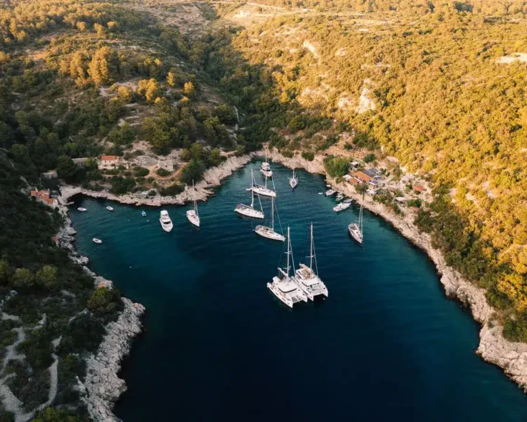 Luftaufnahme einer ruhigen Bucht mit Segelyachten, die im klaren blauen Wasser vertäut sind, umgeben von einer zerklüfteten Küste und dichten grünen Wäldern.