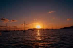 Ein ruhiger Sonnenuntergang über einem Gewässer mit den Silhouetten mehrerer Segelboote, die unter dem leuchtenden Himmel vor Anker liegen und so eine idyllische Segelurlaubsszenerie schaffen.