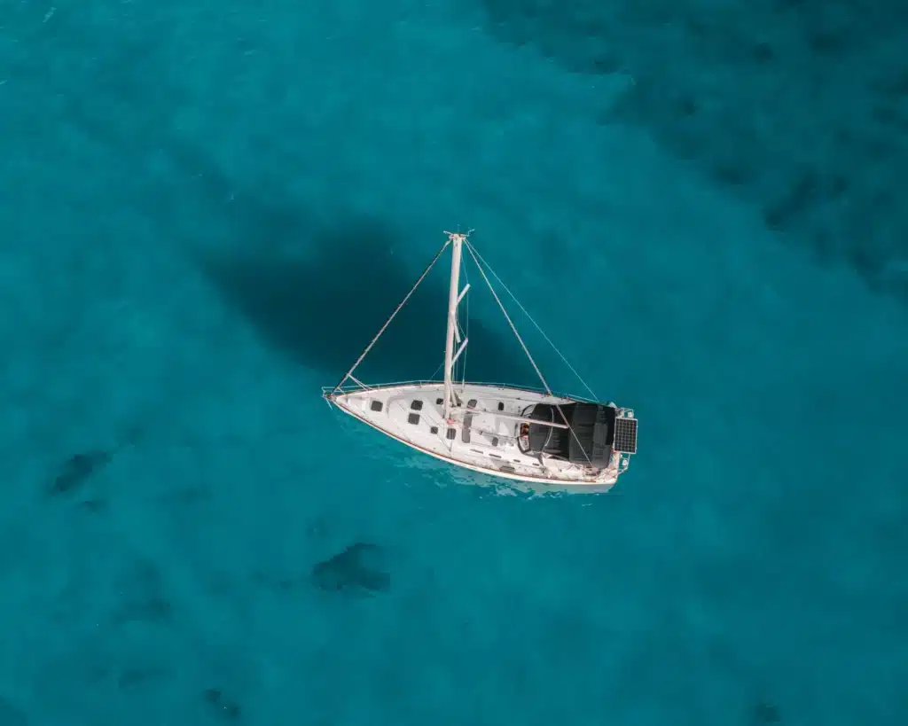 Luftaufnahme eines weißen Segelboots, das auf klarem, türkisfarbenem Wasser schwimmt. Auf dem Segeltörn sind keine Menschen zu sehen, was die Ruhe und Abgeschiedenheit der Umgebung unterstreicht.