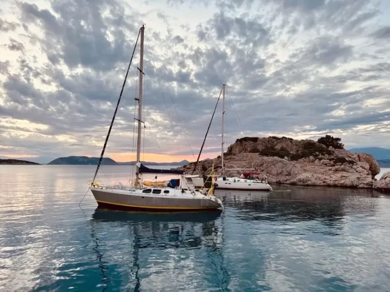 Zwei Segelboote ankern in der Nähe einer felsigen Insel vor der Kulisse eines farbenprächtigen Sonnenuntergangs, der sich während ihres Segeltörns im ruhigen Wasser spiegelt.