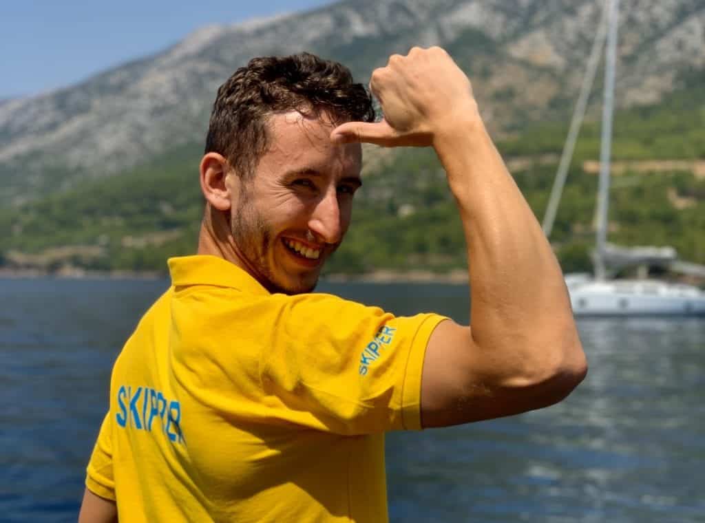 Ein lächelnder Mann in einem gelben „Skipper“-Shirt formt mit der Hand auf der Stirn ein L, im Hintergrund sind ein See und Berge zu sehen, während er im Segelurlaub ist. Skipper werden: Dein Weg zum Kapitänspatent