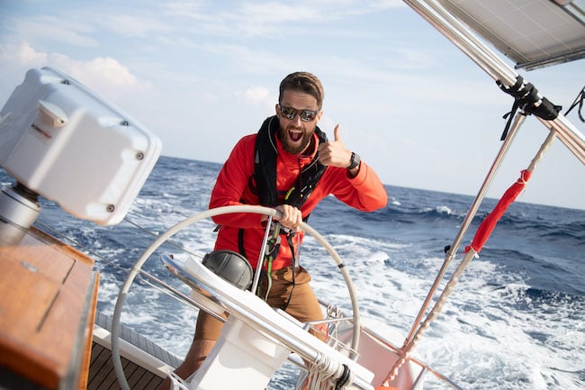 Ein Mann mit Bart, Sonnenbrille und roter Jacke steuert während seiner Segelreise ein Segelboot und streckt den Daumen nach oben, mit dem Meer im Hintergrund.