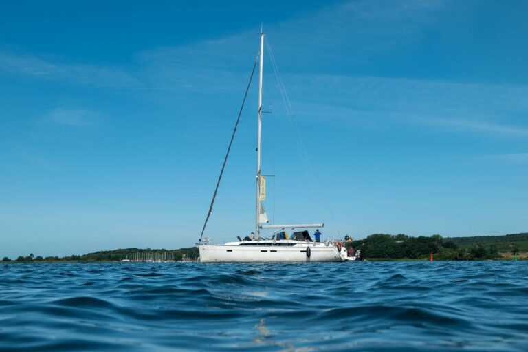 Eine Segelyacht mit einem ungesetzten Segel auf ruhigem blauen Wasser, unter einem klaren Himmel, mit sichtbaren Personen auf dem Achterdeck.
