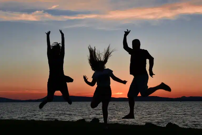 Drei Menschen springen während ihres Segelurlaubs fröhlich am Seeufer vor dem Hintergrund eines Sonnenuntergangs herum und werden von den leuchtenden Farben des Abendhimmels als Silhouetten abgehoben.
