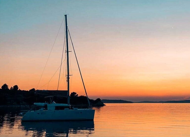 Ein ruhiger Sonnenuntergang mit einem leuchtend orangefarbenen Himmel, der sich im ruhigen Wasser spiegelt und ein Segelboot von Segelreise in der Nähe einer von Bäumen gesäumten Küste hervorhebt.