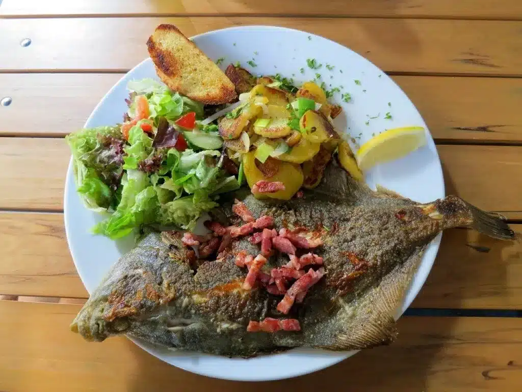 Ein Teller mit einer ganzen gegrillten Forelle, belegt mit knusprigen Speckstücken, dazu Bratkartoffeln, eine Scheibe Knoblauchbrot und ein frischer grüner Salat, serviert auf einem Holztisch während einer