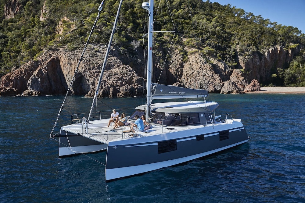 Ein Paar entspannt auf einer Segelyacht und genießt Getränke und Gespräche in der Nähe einer felsigen Küste mit üppigem Grün unter einem klaren blauen Himmel. Luxus und Privatsphäre