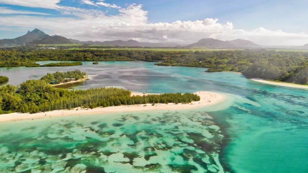 Luftaufnahme einer tropischen Küste mit klarem, türkisfarbenem Wasser, Sandstränden und üppigem Grün, mit Bergen im Hintergrund unter einem bewölkten Himmel, mit einer anmutig segelnden Segelyacht. Seychellen Törn Luxus Abenteuer