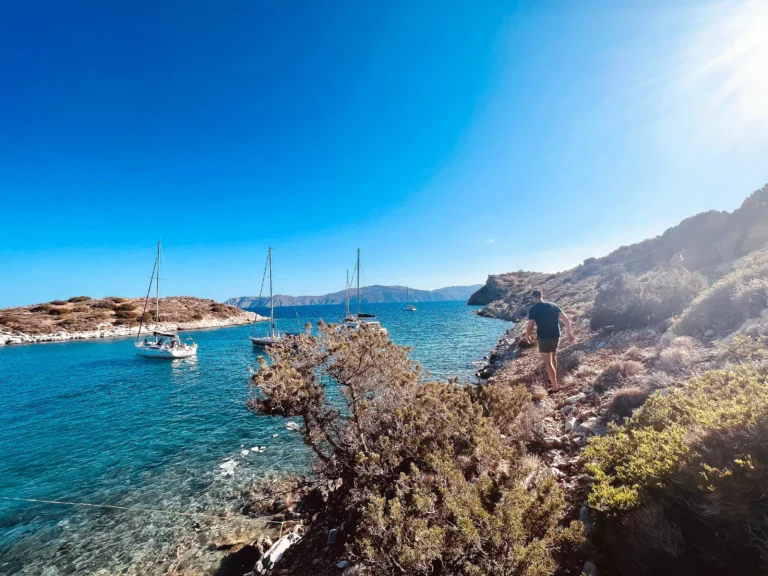 Eine malerische Küstenansicht mit klarem, blauem Wasser und verankerten Segelbooten, darunter ein Katamaran. Felsiges Gelände im Vordergrund, mit einer Person von hinten, die unter einem hellen, sonnigen Himmel auf das Meer blickt.