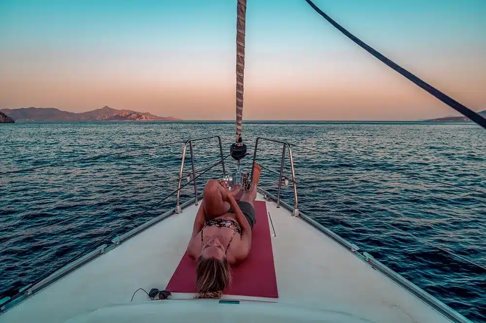 Eine Person entspannt sich während eines Segeltörns auf dem Bug einer Segelyacht und blickt auf das ruhige Meer bei Sonnenuntergang. In der Ferne sind Berge unter einem warmen, pastellfarbenen Himmel zu sehen.