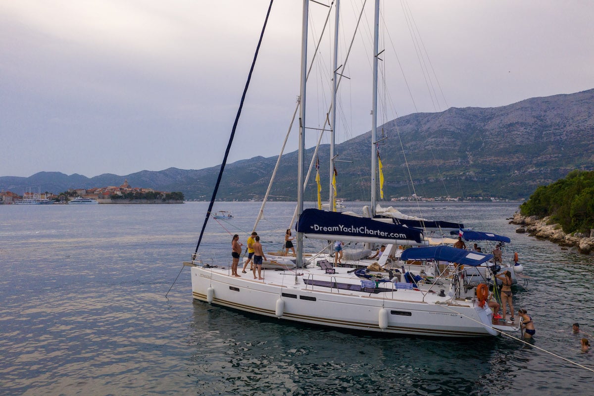 Eine Gruppe von Menschen genießt einen Segeltörn auf einer weißen Segelyacht, die in der Nähe einer üppigen Küste mit Bergen im Hintergrund unter einem bewölkten Himmel vor Anker liegt.