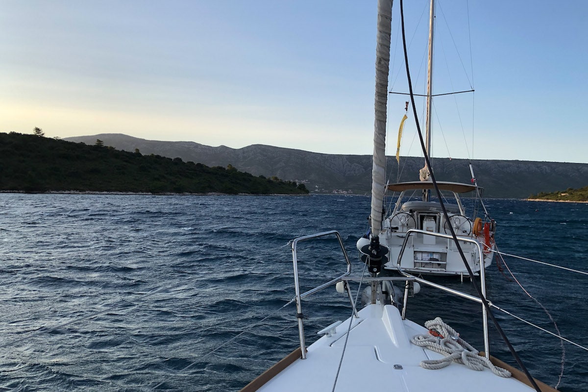 Blick vom Bug einer Segelyacht, die sich durch das Wasser auf eine Küste mit Bergen im Hintergrund zubewegt, unter einem klaren Himmel in der Abenddämmerung.