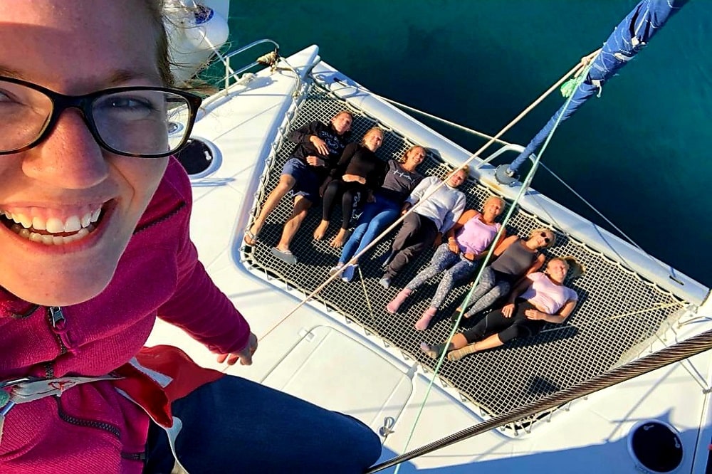 Eine Frau macht ein Selfie und lächelt in die Kamera, während andere hinter ihr auf dem Netztrampolin einer Segelyacht über dem türkisfarbenen Wasser entspannen.