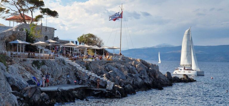 Eine Küstenszene mit Menschen in einem Straßencafé auf einer felsigen Klippe, einer großen kroatischen Flagge, die weht, und einem Segelboot auf einer Segelreise, das auf dem ruhigen Meer unter einem teilweise