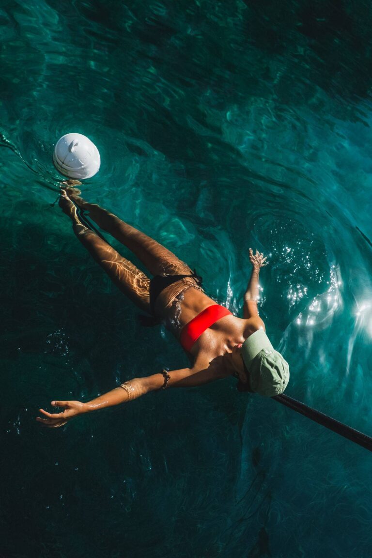 Eine Person in einem roten Bikinioberteil und einer grünen Hose schwimmt während eines Segeltörns im klaren blauen Wasser und versucht, einen weißen Ball zu fangen. Sonnenlicht erzeugt schimmernde Muster auf der Oberfläche.