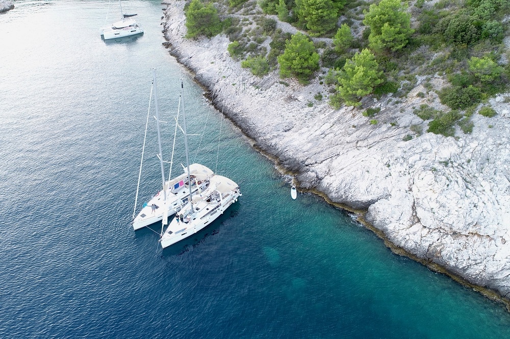 Luftaufnahme einer Segelyacht und eines kleineren Bootes, die in der Nähe einer felsigen Küste mit üppigem Grün im klaren blauen Wasser vor Anker liegen. In der Ferne ist eine weitere Yacht zu sehen.