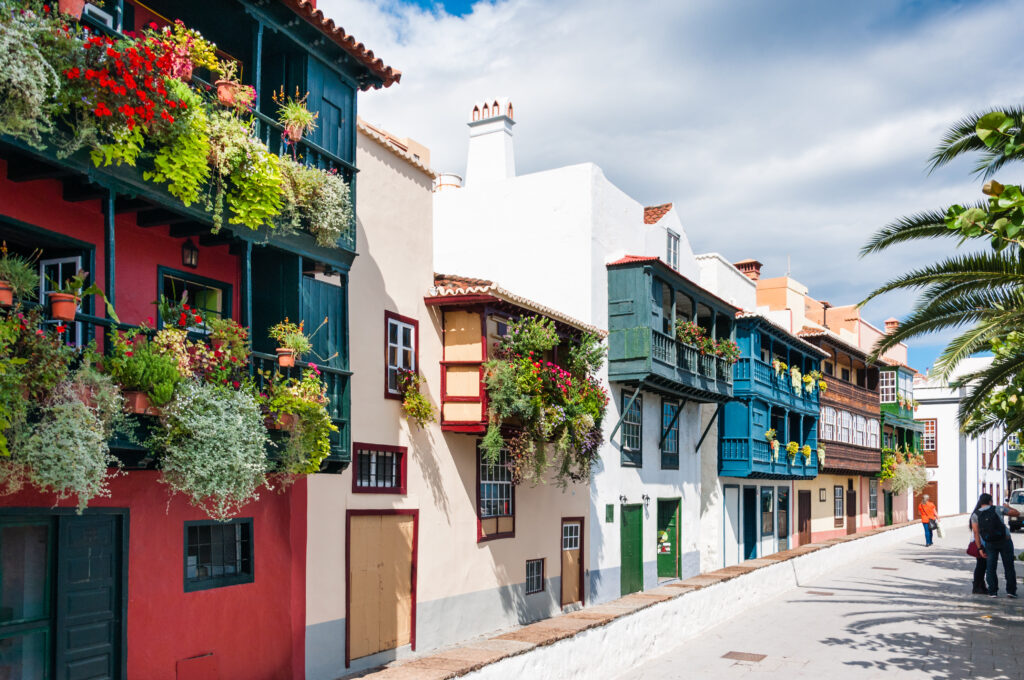 Eine malerische Straße gesäumt von lebendigen, farbenfrohen Häusern, geschmückt mit blühenden Blumenkästen und Balkonen unter einem strahlend blauen Himmel, mit ein paar Menschen, die auf ihre Segelyacht zulaufen.