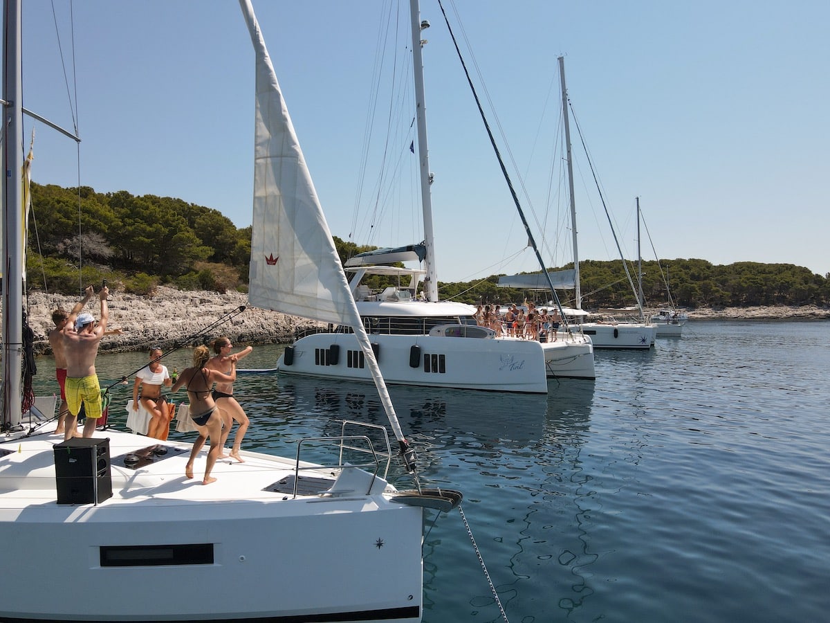 Eine Gruppe von Menschen genießt einen sonnigen Tag auf einem Segelboot in der Nähe einer felsigen Küste. Einige tanzen und winken, im Hintergrund sind bewaldete Hügel zu sehen.