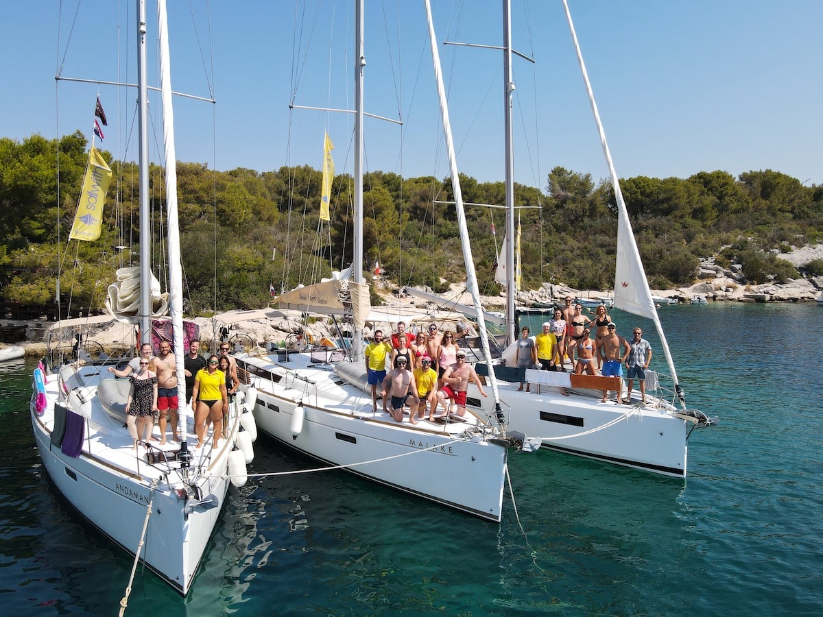 Drei Segelyachten liegen zusammen vor Anker. Auf den Decks stehen Gruppen von Menschen, die einen sonnigen Tag vor der Kulisse von Bäumen und einem klaren blauen Himmel genießen.