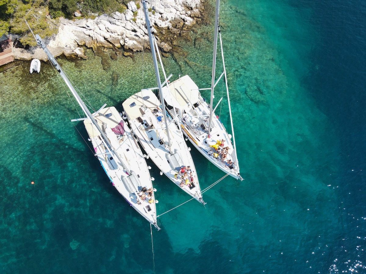 Luftaufnahme von drei Segelbooten, die dicht beieinander in der Nähe einer felsigen Küste mit klarem, türkisfarbenem Wasser vor Anker liegen. Auf den Booten sind Menschen zu sehen, die einen sonnigen Segeltörn-Tag genießen.