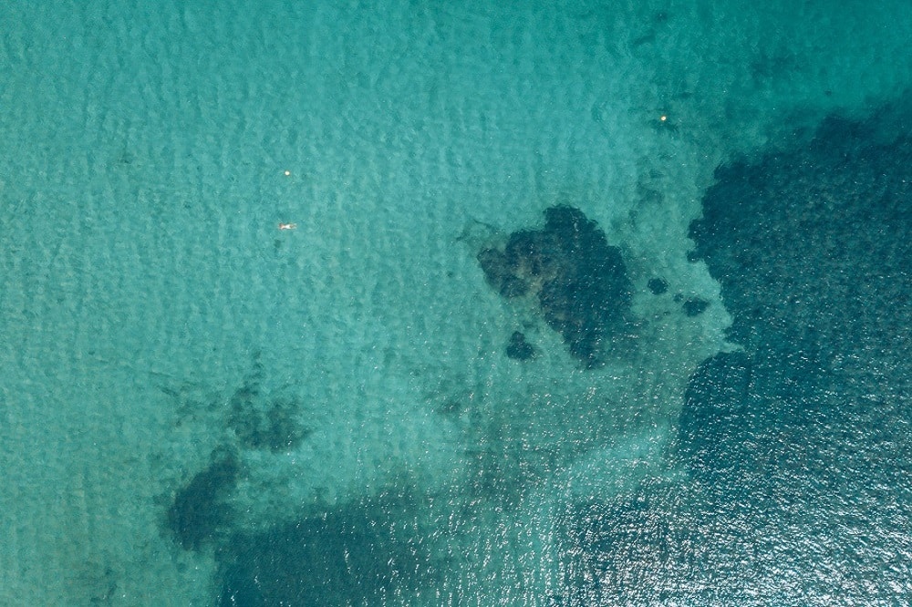 Luftaufnahme eines klaren, türkisfarbenen Meeres mit einer Segelyacht, die durch sanfte Wellen steuert, und sichtbaren dunklen Unterwasserfelsen nahe der Oberfläche.