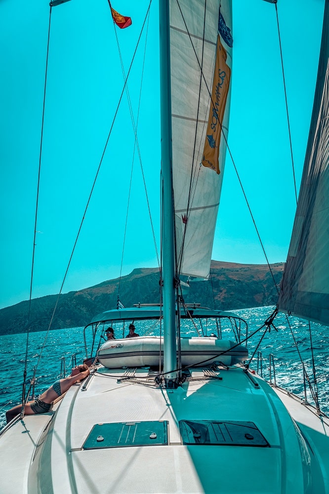 Ein Blick vom Deck einer Segelyacht. Man sieht die gesetzten Segel und die Takelage mit dem Meer und einem Hügel im Hintergrund unter einem strahlend blauen Himmel.