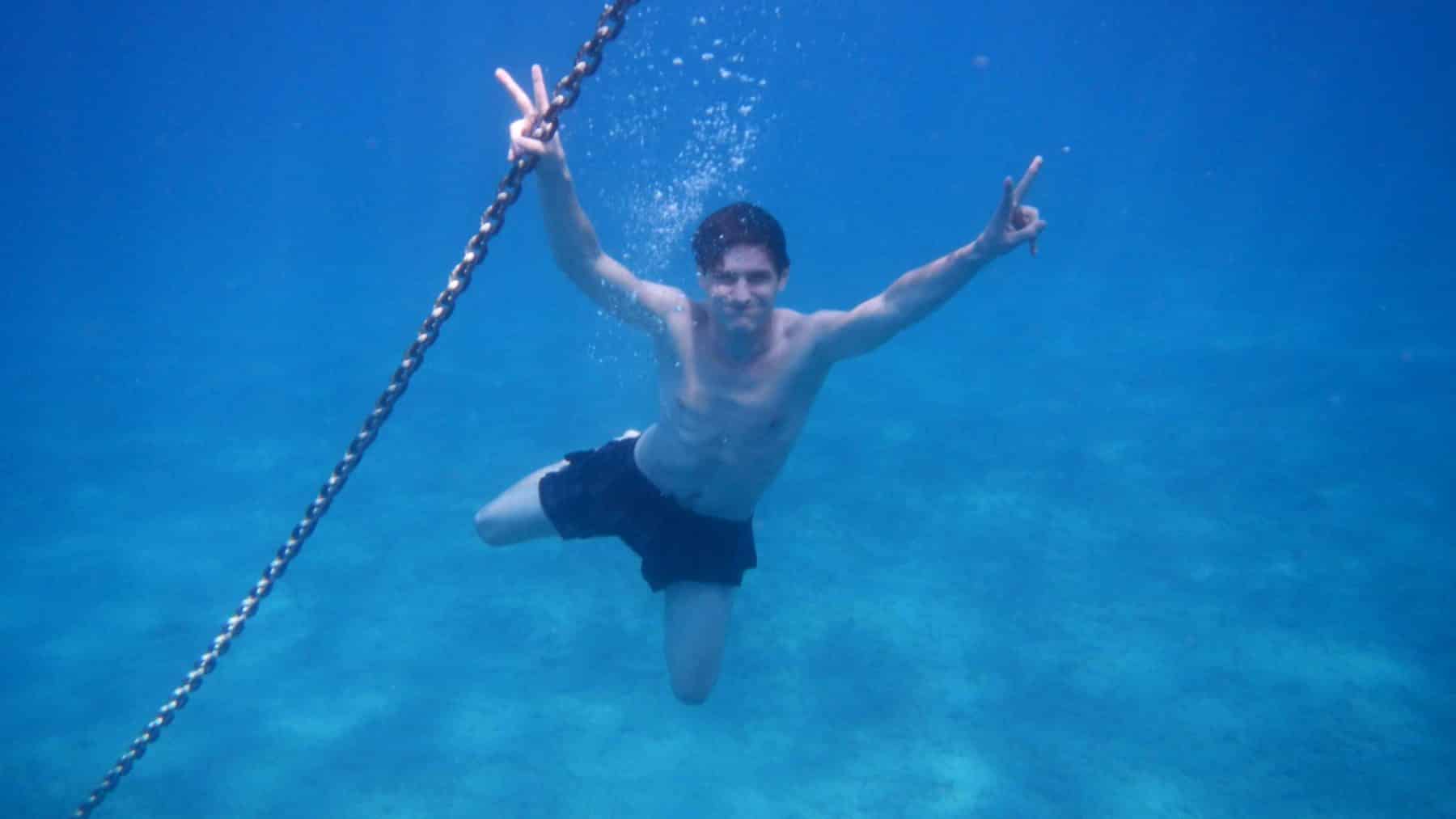 Ein junger Mann macht unter Wasser mit beiden Händen das Peace-Zeichen, während er in die Kamera lächelt und sich an einer Kette festhält, vor der Kulisse des klaren blauen Wassers während eines Segeltörns.