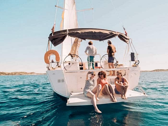 Eine Gruppe von Menschen genießt einen sonnigen Tag auf einer Segelyacht. Einige sitzen am Heck und ein Paar steht am Steuer, umgeben von klarem, blauem Wasser.