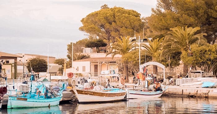 Fischerboote und eine Segelyacht liegen in einem ruhigen Hafen mit mediterraner Architektur und Palmen im Hintergrund, getaucht in warmes, goldenes Licht.