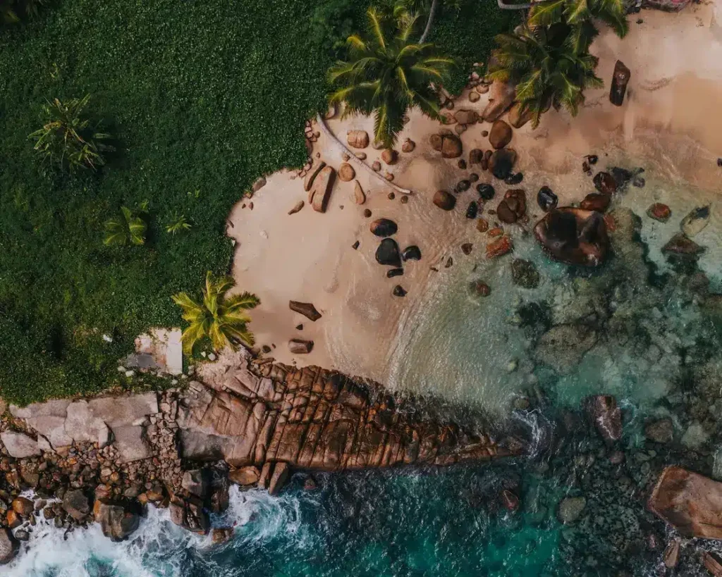 Luftaufnahme eines tropischen Strandes mit klarem türkisfarbenem Wasser, weißem Sand, verstreuten Felsen und üppig grünen Palmen, perfekt für einen Segeltörn. Seychellen Törn Luxus Abenteuer