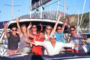 Eine Gruppe von acht jungen Erwachsenen streckt an Bord einer Segelyacht freudig ihre Arme in die Höhe und lächelt unter einem strahlenden Himmel mit Bergen im Hintergrund.