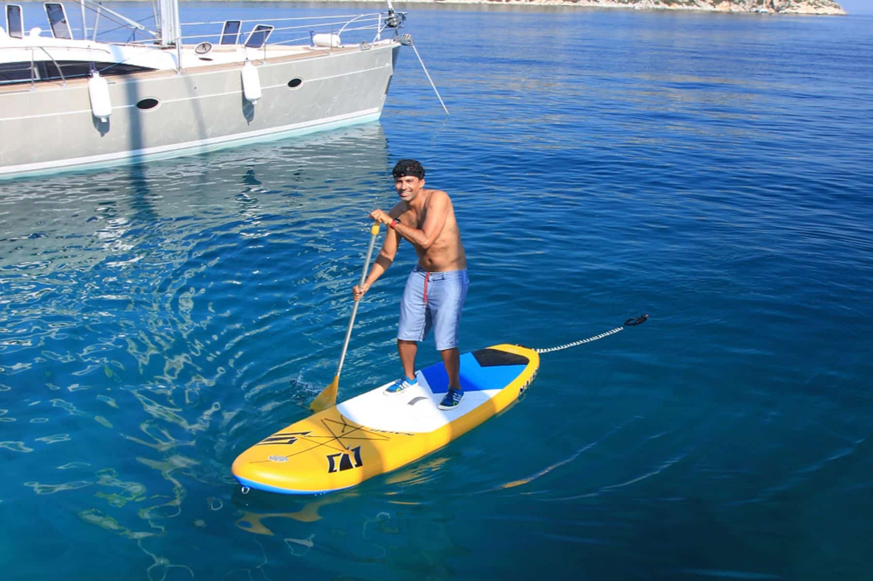 Ein Mann in Badeshorts paddelt auf dem klaren, blauen Wasser neben einer Segelyacht, trägt einen Hut und eine Sonnenbrille und blickt lächelnd in die Kamera.