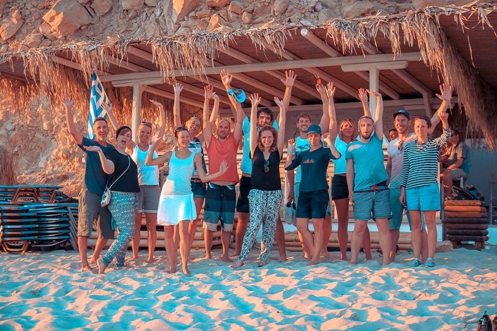 Eine Gruppe fröhlicher Menschen posiert mit erhobenen Händen an einem Sandstrand vor einem rustikalen Gebäude, winkt und lächelt, wobei eine Person nach ihrem Segelausflug eine Flagge hält.