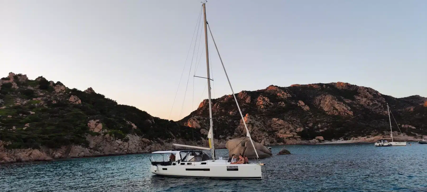 Ein Segelboot schwimmt friedlich in der Nähe felsiger Ufer in einem ruhigen Meer bei Sonnenuntergang, mit anderen Booten in der Ferne und einem rosafarbenen Himmel darüber. Diese idyllische Szene ist perfekt für ein Segelboot.