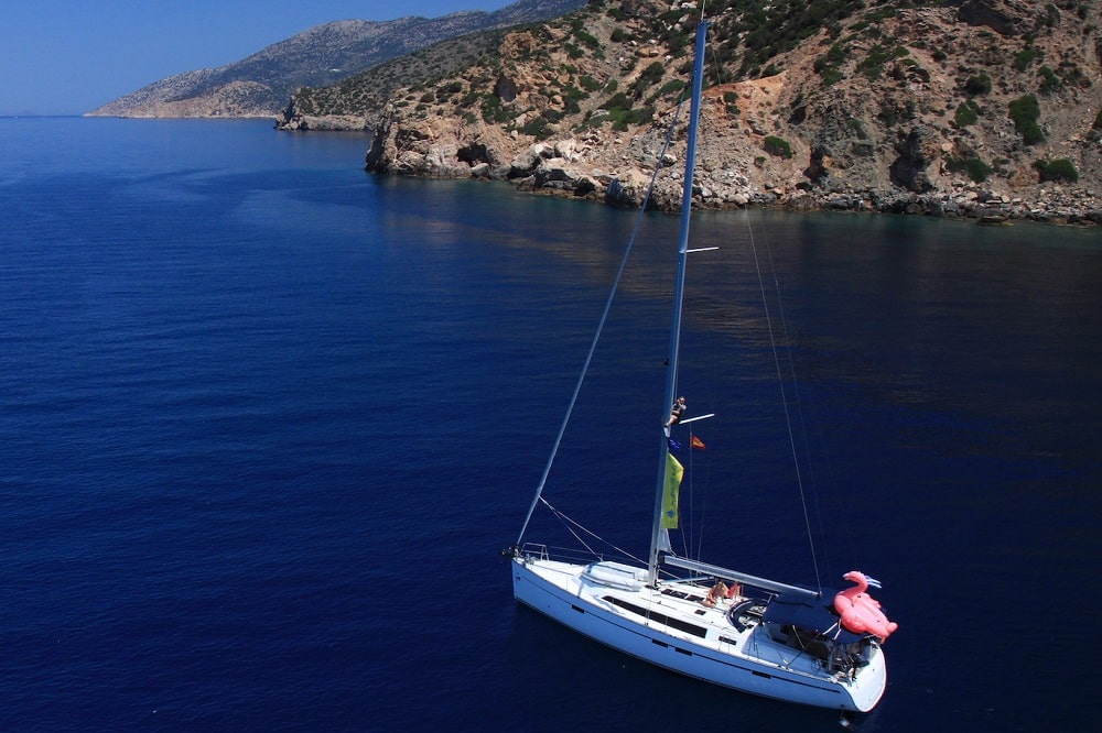 Luftaufnahme einer Segelyacht mit einem leuchtend rosa Spinnaker-Segel, die in der Nähe der felsigen Küste eines klaren blauen Meeres navigiert. Eine Person steht am Bug und gestikuliert.