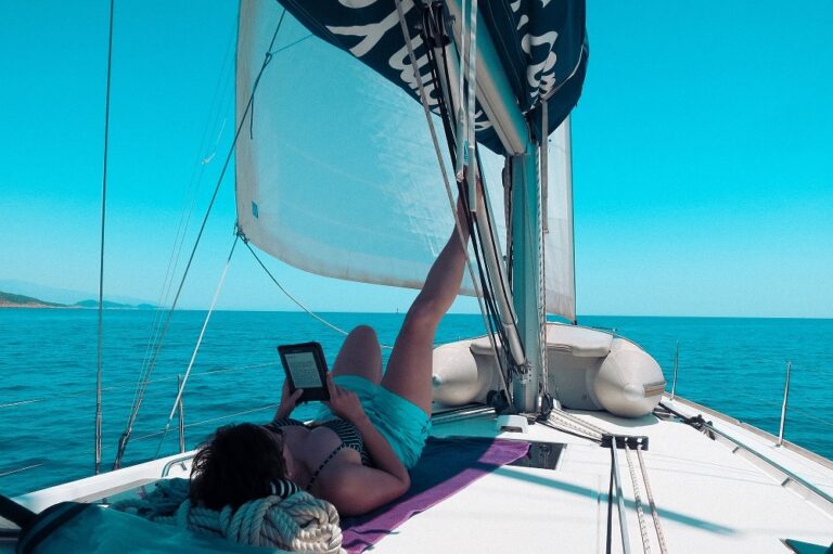Eine Person liegt auf dem Deck einer Segelyacht und liest unter dem Großsegel ein E-Book, im Hintergrund ein klarer blauer Himmel und eine ruhige See.