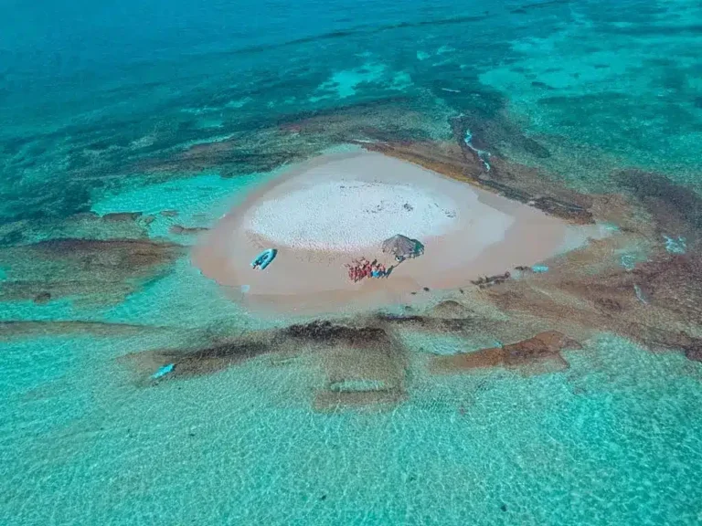 Luftaufnahme einer kleinen, herzförmigen Sandinsel, umgeben von klarem, türkisfarbenem Wasser. Ein paar Menschen und Boote sind zu sehen, perfekt für einen ruhigen Segelurlaub.